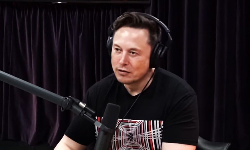 Elon Musk sitzt vor einem Mikrofon und spricht hinein. Er trägt Kopfhörer.