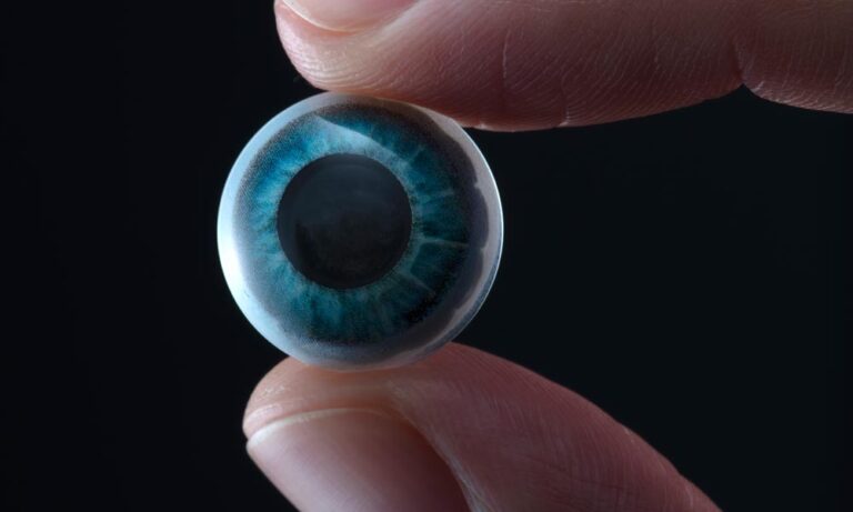 Eine AR-Kontaktlinse wird zwischen zwei Fingern groß vor die Kamera gehalten.