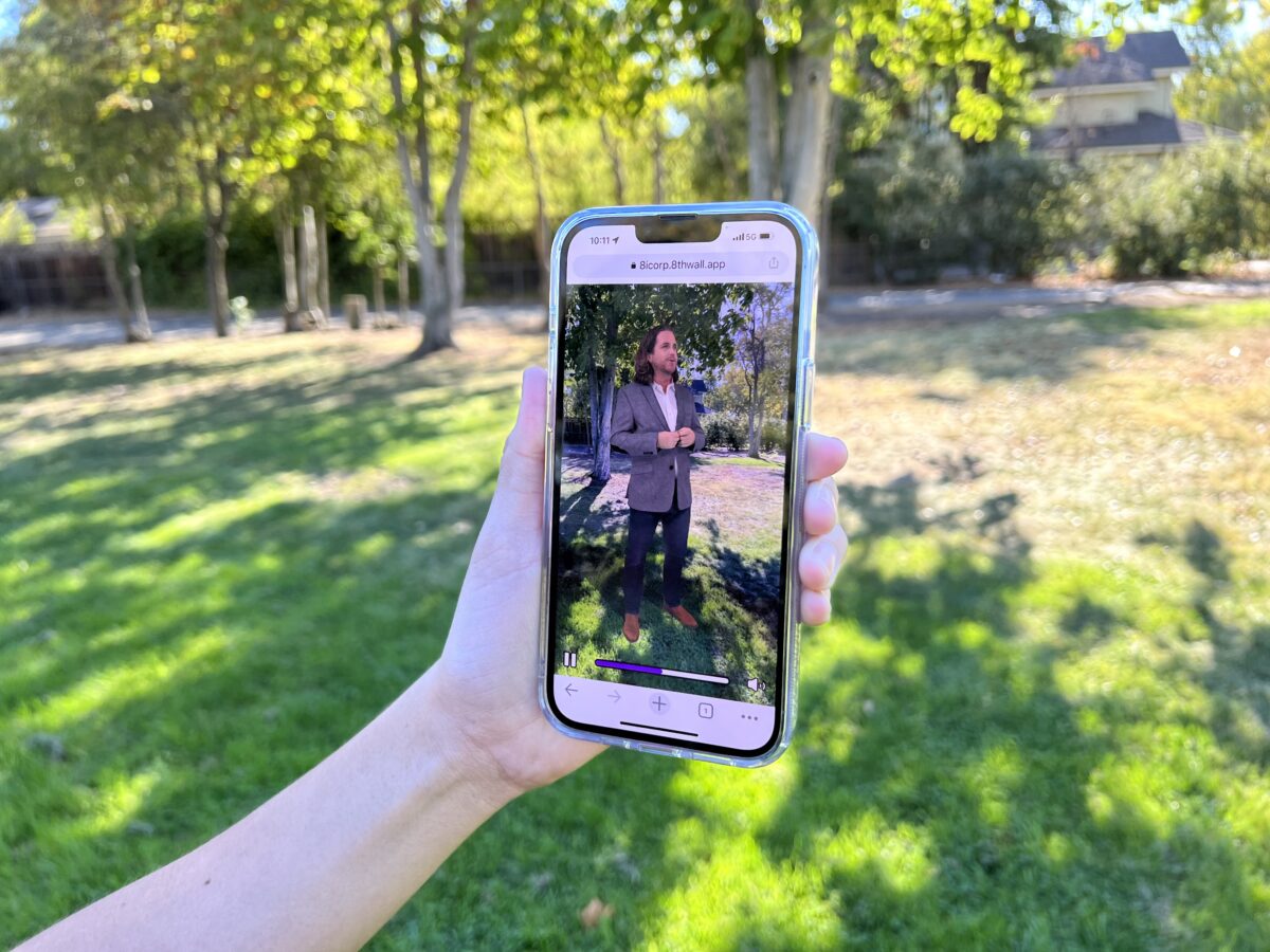 Eine Person hält ein Smartphone auf eine Wiese, auf der Wiese sieht man ein Hologramm eines Mannes stehen, das im Smartphone-Display sichtbar wird.
