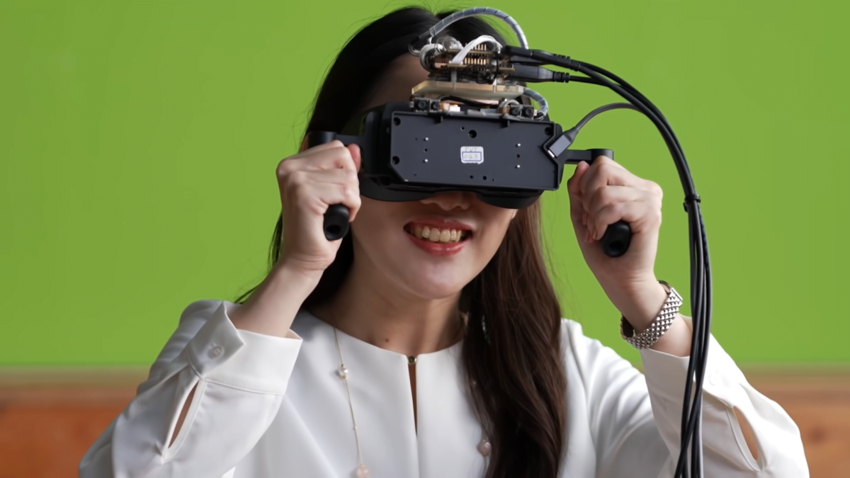 Eine Frau hält sich eine prototypische VR-Brille an zwei Halterungen vors Gesicht. Die VR-Brille hat viele Kabel. Die Frau lächelt.