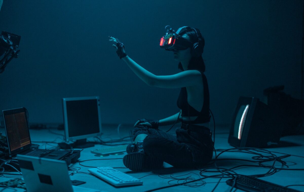 Junge Frau mit VR-Brille sitzt auf dem Boden, umgeben von Computer, und interagiert mit der Hand in einer virtuellen Welt