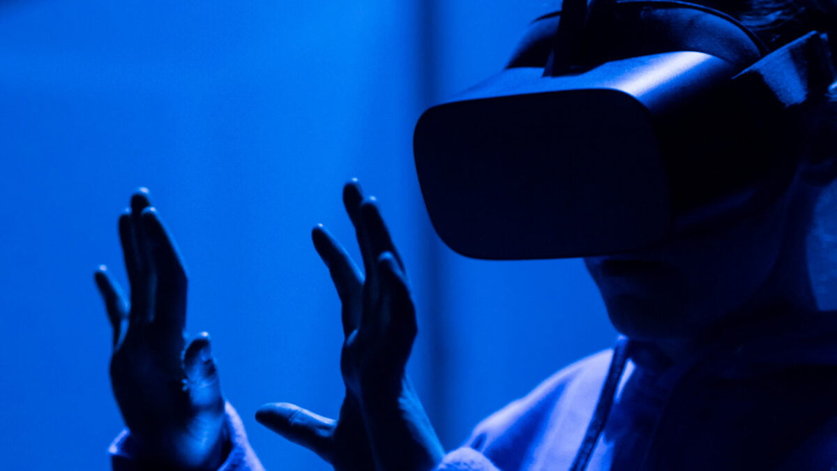 Frau mit VR-Brille interagiert mit ihren Händen in der virtuellen Welt, steht selbst im Schatten eines blau beleuchteten, dunklen Raums des Staatstheaters Augsburg