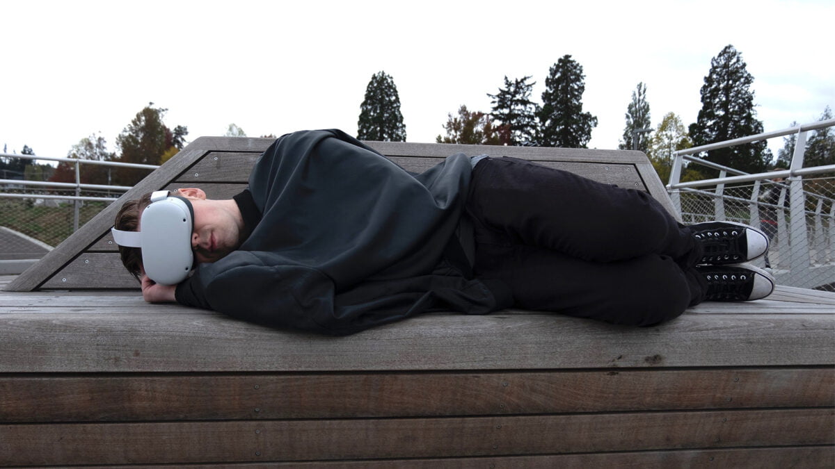 Ein junger Mann liegt zugedeckt auf einer Bank im Freien mit Meta Quest 2 im Gesicht.