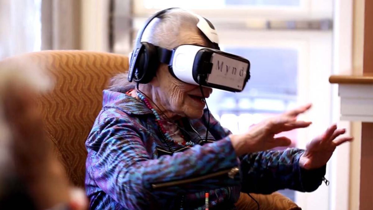 Eine Seniorin sitzt mit einer VR-Brille auf dem Kopf auf einem Sessel und streckt die Arme nach vorn aus.