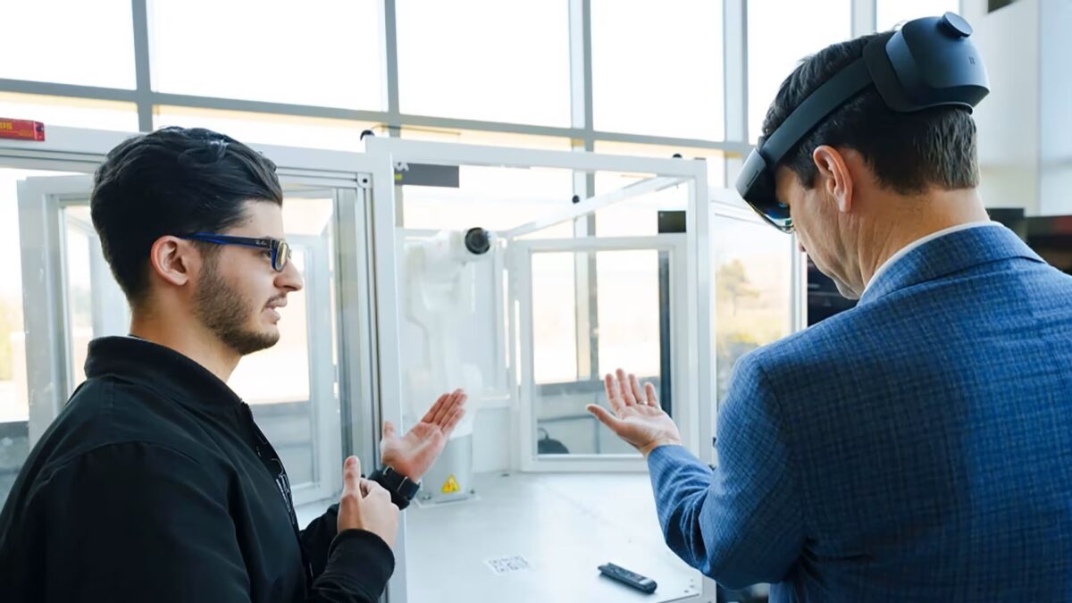 Zwei Männer stehen nebeneinander, der rechte hat eine HoloLens 2 auf dem Kopf, während der linke ihm die Gestensteuerung erklärt