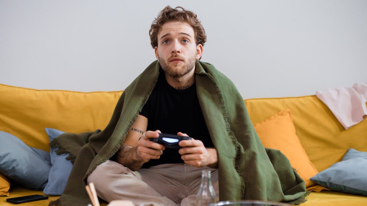 Ein junger Mann sitzt auf einer Couch in eine Decke eingehüllt und hält eine Gamepad in der Hand.
