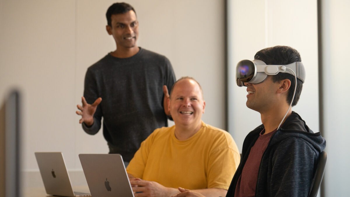 Ein Entwickler mit Apple Vision Pro im Gesicht lächelt. Neben ihm stehen zwei Apple-Angestellte, die die Reaktion beobachten.