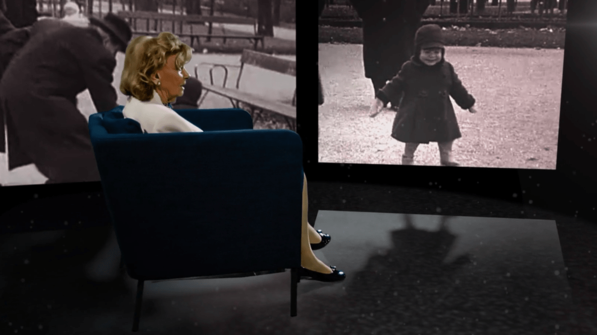 Eine alte Frau sitzt auf einem Sessel während im Hintergrund Aufnahmen ihrer Kindheit auf einer Leinwand laufen