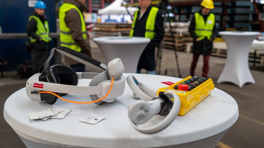 Auf einem Tisch in einer Industriehalle liegen eine VR-Brille und eine Kranfernbedienung, an der ein VR-Controller befestigt ist.