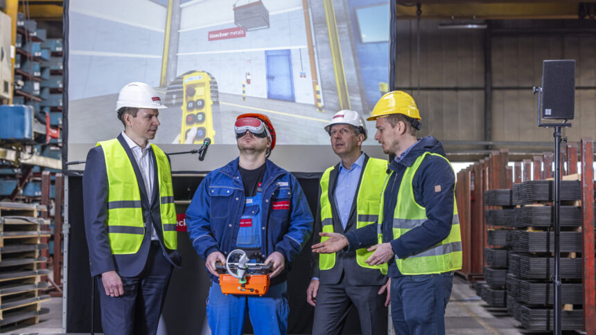 In einer Industriehalle stehen vier Männer in Sicherheitskleidung. Einer davon trägt eine VR-Brille und bedient einen virtuellen Kran. 